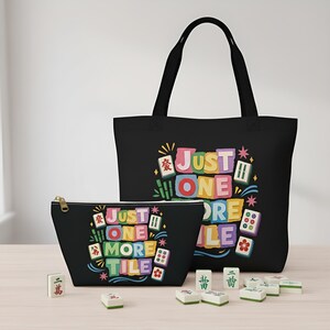 May include: Black tote bag and zippered pouch featuring the text "Just One More Tile" in colorful block letters, with Mahjong tile illustrations. Several Mahjong tiles are scattered in front of the bags.