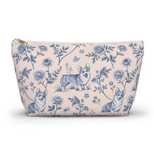May include: A zippered pouch with a light pink background and a repeating pattern of blue corgis, flowers, and foliage. The pouch has a gold-colored zipper and is designed to hold various items.