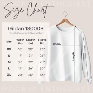 May include: A white youth crewneck sweatshirt hanging on a wooden hanger. The image includes a size chart with measurements in inches for sizes XS to XL. The chart details width, length, and sleeve measurements for the Gildan 18000B sweatshirt.