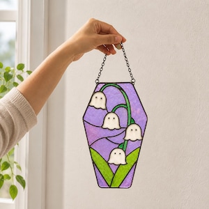May include: A coffin-shaped stained glass suncatcher featuring four white ghost-like lily of the valley flowers with black dot eyes, green stems and leaves, and a textured purple background, suspended by a black metal chain for hanging display.