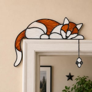 Könnte beinhalten: Ein Buntglas-Katzenornament, das auf einem Türrahmen aufliegt. Die Katze besteht aus orangefarbenen und weißen, strukturierten Glaspaneelen mit schwarzen Bleilinien. Ein kleiner weißer Maus-Anhänger hängt an einer schwarzen Metallkette.
