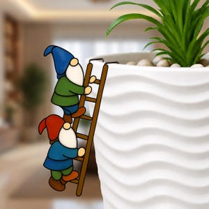 May include: Two garden gnomes climbing a brown wooden ladder attached to a white ceramic planter. The gnomes wear blue, green, and red outfits with white beards and hats. A green plant with small leaves sits inside the planter.