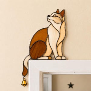 Könnte beinhalten: Eine Buntglas-Katzenfigur in Creme-, Beige- und Brauntönen mit dunklen Bleilinien und einer kleinen gelben Glocke, die an der Schwanzspitze hängt. Die Katze ist in sitzender Position dargestellt und so gestaltet, dass sie auf einem Rahmen sitzt.