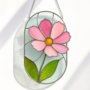 Può includere: Un acchiappasole ovale in vetro colorato raffigurante un fiore rosa con un centro giallo e due foglie verdi. Il fiore è posto su uno sfondo verde chiaro testurizzato, il tutto racchiuso in una cornice di metallo scuro con catena.