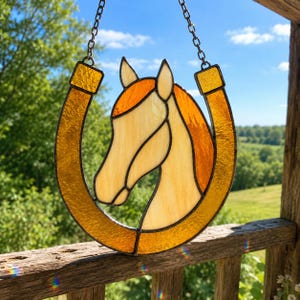 Può includere: Un acchiappasole in vetro colorato raffigurante il profilo di una testa di cavallo all'interno di una cornice a ferro di cavallo. Il cavallo è realizzato in vetro color crema e ambra, mentre il ferro di cavallo è in vetro arancione testurizzato.