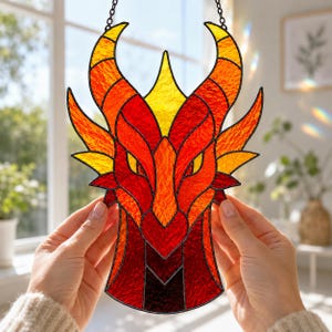 Op de afbeelding: Een glas-in-lood zonnevanger in de vorm van een drakenkop met levendige rode, oranje en gele getextureerde glaspanelen verbonden door donkere metalen soldeerlijnen. Het ontwerp bevat puntige hoorns en gele ogen.