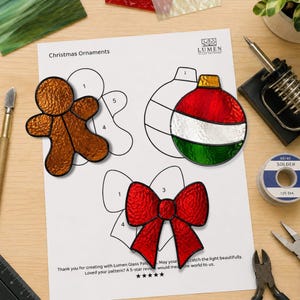 May include: A paper pattern sheet titled Christmas Ornaments by Lumen Glass Patterns featuring three stained glass pieces: a textured brown gingerbread man, a red, white, and green striped bauble, and a bright red bow with a textured finish.