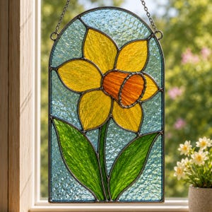 May include: An arched stained glass suncatcher featuring a yellow daffodil with an orange centre and green leaves against a textured light blue background. The piece is framed in dark metal and hangs from a thin silver chain in front of a window.