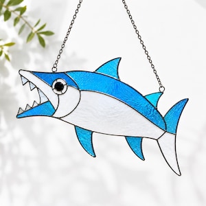 May include: A stained glass suncatcher shaped like a fish with an open mouth and sharp teeth. The fish features a combination of textured blue and opaque white glass panels held together by dark metal solder lines, suspended by a thin metal chain.