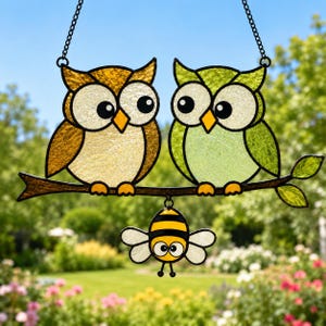 May include: A stained glass suncatcher featuring two owls perched on a branch, one in amber and one in lime green, with a small bumblebee hanging below. The owls have large white eyes and black pupils, while the bee has yellow and black stripes.