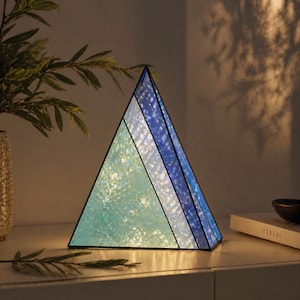 May include: A triangular stained glass lamp featuring three distinct panels in shades of light blue, white, and dark blue. The lamp is illuminated from within, casting a soft glow through the textured glass surfaces framed by thin black metal lines.