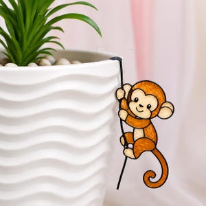 May include: A decorative brown monkey climbing a black line, attached to a white wavy-textured planter. The planter holds a green plant with small white stones at the base. The background is a soft pink.