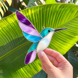 May include: A stained glass hummingbird suncatcher featuring vibrant purple, lavender, teal, and white glass segments held together by thin silver metal solder lines. The bird has a long, pointed black beak and a small circular metal hanging loop.