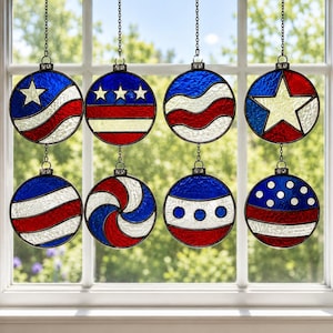 Puede incluir: Ocho adornos redondos de vidrieras con diseños patrióticos en rojo, blanco y azul cuelgan frente a una ventana. Cada adorno presenta un diseño diferente, incluyendo estrellas, rayas y espirales, suspendidos por una cadena.