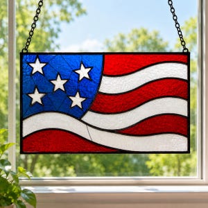 May include: Stained glass panel depicting the American flag, featuring a blue field with white stars and red and white stripes. The panel is framed in black and hangs from a black chain, perfect for patriotic decor.