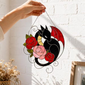 Può includere: Un acchiappasole circolare in vetro colorato raffigurante un drago nero con ventre giallo e ali rosse, circondato da due rose rosse e una rosa rosa chiaro con foglie verdi. L'oggetto è sospeso tramite una sottile catena in metallo argentato.
