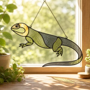 May include: A stained glass lizard suncatcher featuring shades of light green, olive, and translucent grey glass. The lizard is outlined in black metal solder and hangs from a thin metal chain, displaying a curved tail and four distinct legs.
