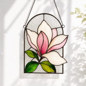May include: A stained glass suncatcher featuring a magnolia flower with white and pink petals and green leaves. The flower is set within an arched frame of clear textured glass, held by a black metal chain for hanging against a window.
