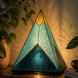Op de afbeelding: Een piramidevormige glas-in-loodlamp met een bergachtig ontwerp. Het bovenste gedeelte is amberkleurig, terwijl het midden en de basis bestaan uit verschillende tinten getextureerd blauw en groenblauw glas, bijeengehouden door een donker metalen frame.
