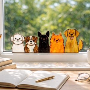 May include: A row of five dog-shaped decorations in various breeds and colors, including a white Maltese, a brown and white Bulldog, a black French Bulldog, an orange Pomeranian, and a yellow Golden Retriever. They are displayed on a windowsill.