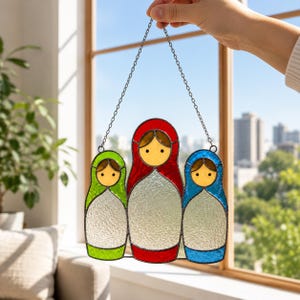 Op de afbeelding: Een glas-in-lood suncatcher met drie matroesjka-poppen op een rij. De linkerfiguur is groen, de middelste rood en de rechter blauw. Elke pop heeft een geel gezicht en een wit lichaam met textuur, verbonden door een metalen ketting.