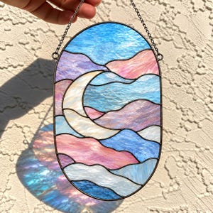 May include: An oval stained glass suncatcher featuring a crescent moon set against a background of wavy, textured glass panels in shades of blue, pink, and white. The piece is framed in dark metal and hangs from a thin black chain.