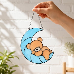Può includere: Un acchiappasole in vetro colorato raffigurante una luna crescente azzurra con un orso color marrone chiaro che dorme sopra di essa. L'oggetto è realizzato con linee di saldatura in metallo scuro e include una sottile catena metallica per appenderlo.