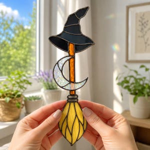 May include: A stained glass suncatcher featuring a black witch hat at the top, a crescent moon with an iridescent texture in the center, and a yellow broomstick at the bottom, all connected by a vertical orange glass rod with dark metal solder.