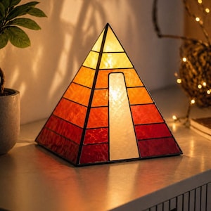 May include: A pyramid-shaped stained glass lamp featuring horizontal panels in a gradient of yellow, orange, and red hues. The structure includes a vertical white rectangular panel on the front face, all held together by dark metal soldering lines.