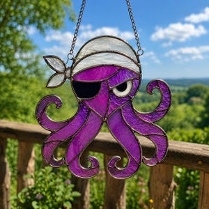 Peut inclure: Un attrape-soleil en vitrail en forme de pieuvre violette portant un bandana de pirate blanc et un cache-œil noir. La pieuvre possède huit tentacules recourbés et est suspendue par une chaîne en métal argenté sur un fond naturel extérieur.