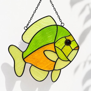 May include: A stained glass suncatcher shaped like a fish, featuring vibrant segments of lime green, bright orange, and pale yellow glass. The piece is outlined in black metal solder and hangs from a black metal chain for display in a window.