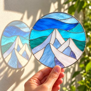 May include: A circular stained glass suncatcher featuring a mountain landscape with white, light blue, and dark blue glass segments. The design includes layered peaks and a wavy sky pattern, all held together by thin metal solder lines in a round frame.