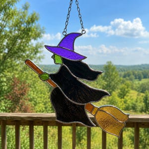 May include: A stained glass suncatcher depicting a witch in flight on a broomstick. The witch wears a purple pointed hat and a black robe, with a green face and hands. The broomstick features an orange handle and a textured yellow brush.