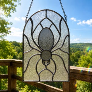 May include: An arched stained glass suncatcher featuring a central spider design. The piece is constructed with textured clear glass panels held together by dark metal lead lines. It hangs from a thin metal chain, displaying a spider silhouette.