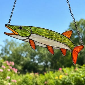May include: A stained glass suncatcher shaped like a fish with a green textured body, a white underbelly, and vibrant orange fins and tail. The piece is suspended by a silver metal chain, featuring dark lead lines outlining the glass segments.