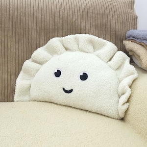 Peut inclure: Un coussin en peluche semi-circulaire en tissu sherpa texturé blanc cassé. Le coussin a la forme d'un ravioli avec un bord froncé sur le dessus. Il présente deux yeux circulaires noirs et une petite ligne noire incurvée pour un sourire.