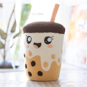 May include: A plush boba tea toy featuring a tan base with dark brown tapioca pearls, a white cream top, and a dark brown lid. The cup has a smiling face with large black eyes, eyelashes, and pink cheeks, topped with a light peach-coloured straw.