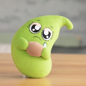 May include: A plush, green, bean-shaped character with large, expressive eyes and a sad expression. The character is holding a small, brown object and has a single tear. The toy is on a wooden surface.