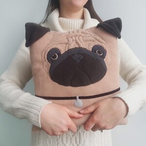 Puede incluir: Un coj&iacute;n en forma de pug de color beige con nariz, orejas y collar negros. El coj&iacute;n tiene detalles bordados para los ojos y una peque&ntilde;a etiqueta. El art&iacute;culo es un objeto decorativo.
