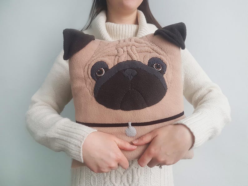 Puede incluir: Un coj&iacute;n en forma de pug de color beige con nariz, orejas y collar negros. El coj&iacute;n tiene detalles bordados para los ojos y una peque&ntilde;a etiqueta. El art&iacute;culo es un objeto decorativo.