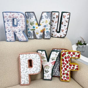 Puede incluir: Letras decorativas de tela con diversos patrones: conejitos, pulpos, ardillas, dinosaurios y fresas. Las letras, delineadas en colores a juego, están dispuestas sobre un sofá beige. Las letras deletrean "RMU" y "PYF". Cada letra mide unos 20 cm de alto.
