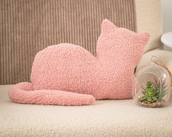 May include: A pink cat-shaped pillow made of textured sherpa fabric, featuring a long tail and pointed ears. Beside the pillow sits a clear glass terrarium containing a small green succulent plant and decorative pebbles on a light surface.