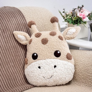 Può includere: Un cuscino decorativo in peluche a forma di testa di giraffa, realizzato in tessuto sherpa beige e bianco. Il cuscino presenta macchie ricamate marroni, due piccole corna, orecchie a punta, occhi rotondi marrone scuro e una bocca sorridente.