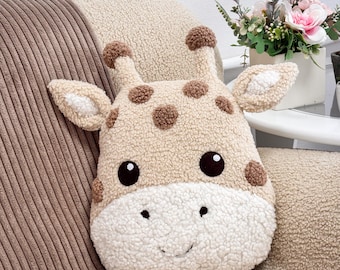 May include: A plush throw pillow shaped like a giraffe head, crafted from beige and white sherpa fabric. The pillow features brown embroidered spots, two small horns, pointed ears, dark brown circular eyes, and a stitched smiling mouth.