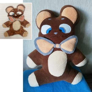 Può includere: Un peluche orsetto marrone fatto a mano con un papillon marrone chiaro e accenti bianchi sulle zampe e sulla pancia. L'orso ha occhi blu e orecchie marrone chiaro. Un disegno dell'orso è nell'angolo in alto a sinistra.