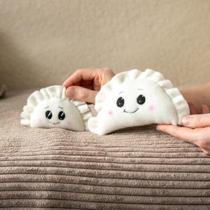 May include: Two white plush dumplings with embroidered faces, including black eyes and pink cheeks. One dumpling is held by hands, while the other rests on a brown, textured surface. The dumplings have a ruffled edge.