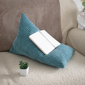 May include: A teal corduroy wedge pillow with a textured ribbed surface, featuring a white spiral-bound notebook resting on its angled top face, accompanied by a small white square planter containing a green succulent plant.
