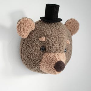 Può includere: Una decorazione da parete a forma di testa di orso in peluche, realizzata in tessuto pile testurizzato marrone e beige. L'orso ha due occhi in plastica scura, un naso marrone scuro e indossa un piccolo cilindro in feltro nero sulla testa.