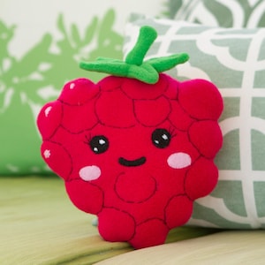 May include: A plush, red raspberry with a smiling face, black eyes, and pink cheeks. The raspberry has a green stem and leaves. The background includes green and white patterned pillows.