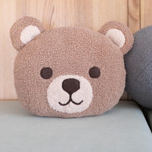 Könnte beinhalten: Ein kuscheliges, teddybärförmiges Kissen in Brauntönen, mit weißer Schnauze und Innenohren. Der Bär hat dunkelbraune Augen und ein aufgesticktes Lächeln. Das Kissen liegt auf einer hellblauen Oberfläche, mit einem Holz Hintergrund.